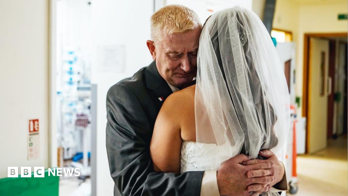 Sinèad Jackson has her back to the camera in a white veil and strapless white gown. She has dark brown hair. Leslie wears a dark suit. He has short white hair.