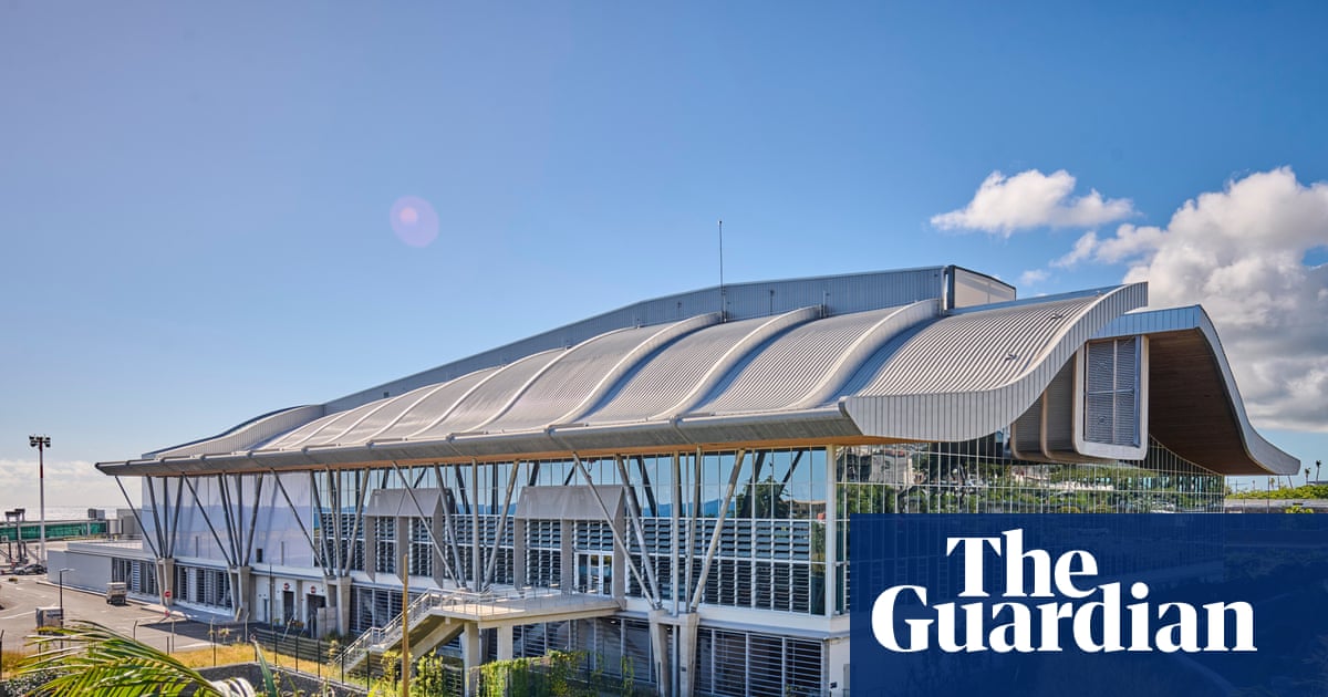 Exterior of the arrivals terminal, which has a curved roof and glass facade. Plants are growing in the foreground surrounding the building